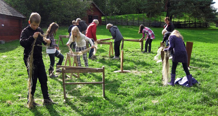 Ett tiotal skolelever bråkar och skäktar lin framför en rödmålad timrad linbastu.