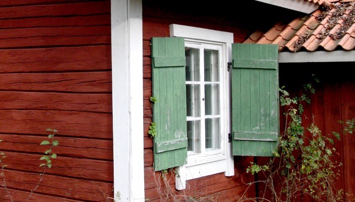 Detalj av falurött torp med spröjsat fönster och gröna fönsterluckor
