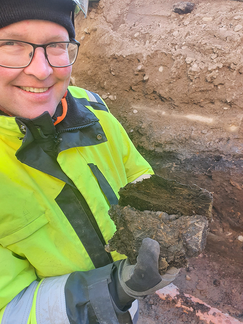 En leende arkeolog i varselkläder håller upp brun jord som innehåller små träbitar och halm 