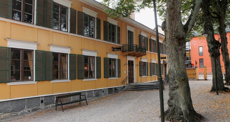 Gult tvåvåningshus med stående träpanel och gröna fönsterluckor.