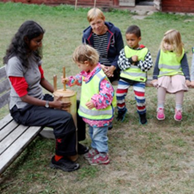 Ett förskolebarn kärnar smör, två barn och två pedagoger tittar på.