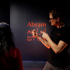 En man pekar på namnet Abram i rött, i latinsk, arabisk och hebreisk skrift på en mörk vägg i en utställning.