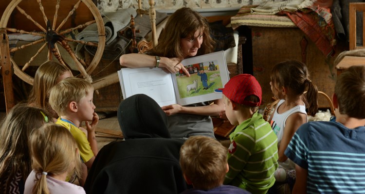 Barn lyssnar när en pedagog läser boken Pelles Nya kläder bredvid en spinnrock.