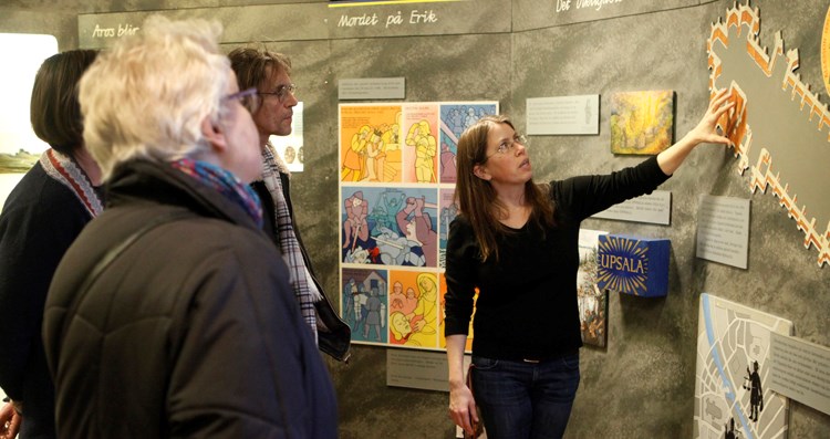 Tre personer lyssnar på en museipedagog som pekar på en ritning över Uppsala domkyrka.